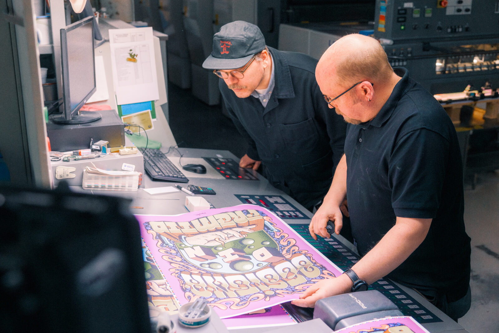 Två män granskar ett färgglatt tryck på ett kontor med datorer och arbetsmaterial.
