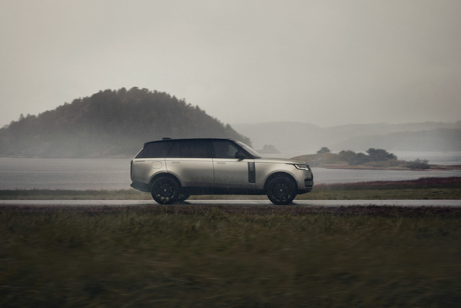 Lyxig SUV kör längs en väg vid en sjö i regnigt landskap.